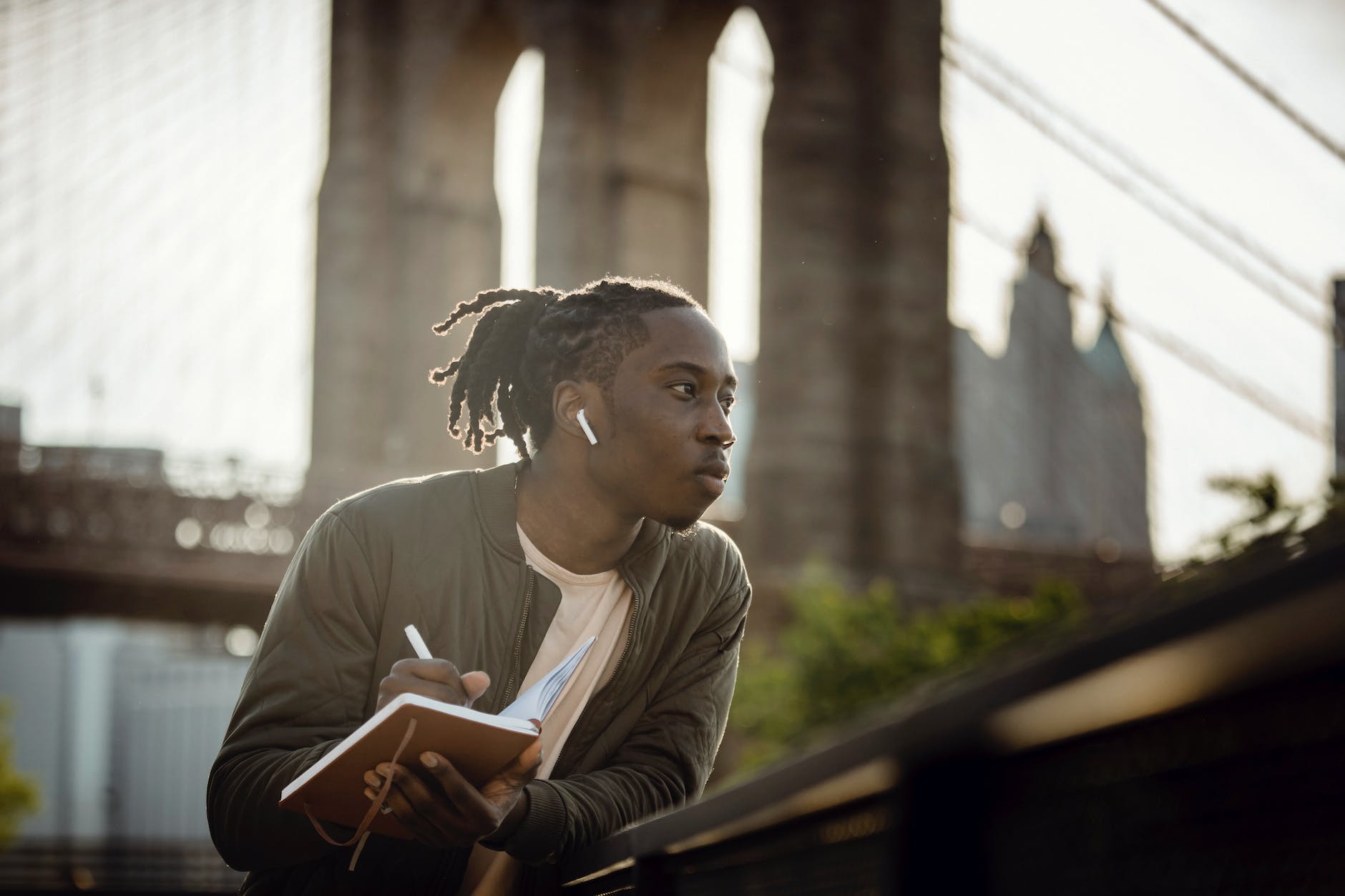 positive black man listening to music with wireless earphones and taking notes in diary on street